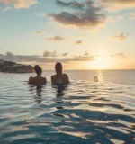 Two people relax in an infinity pool at a luxury seaside resort, with a stunning ocean sunset horizon in view