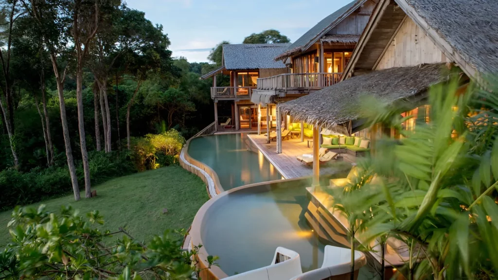 A tropical resort featuring a pool and a thatched roof, surrounded by lush greenery.